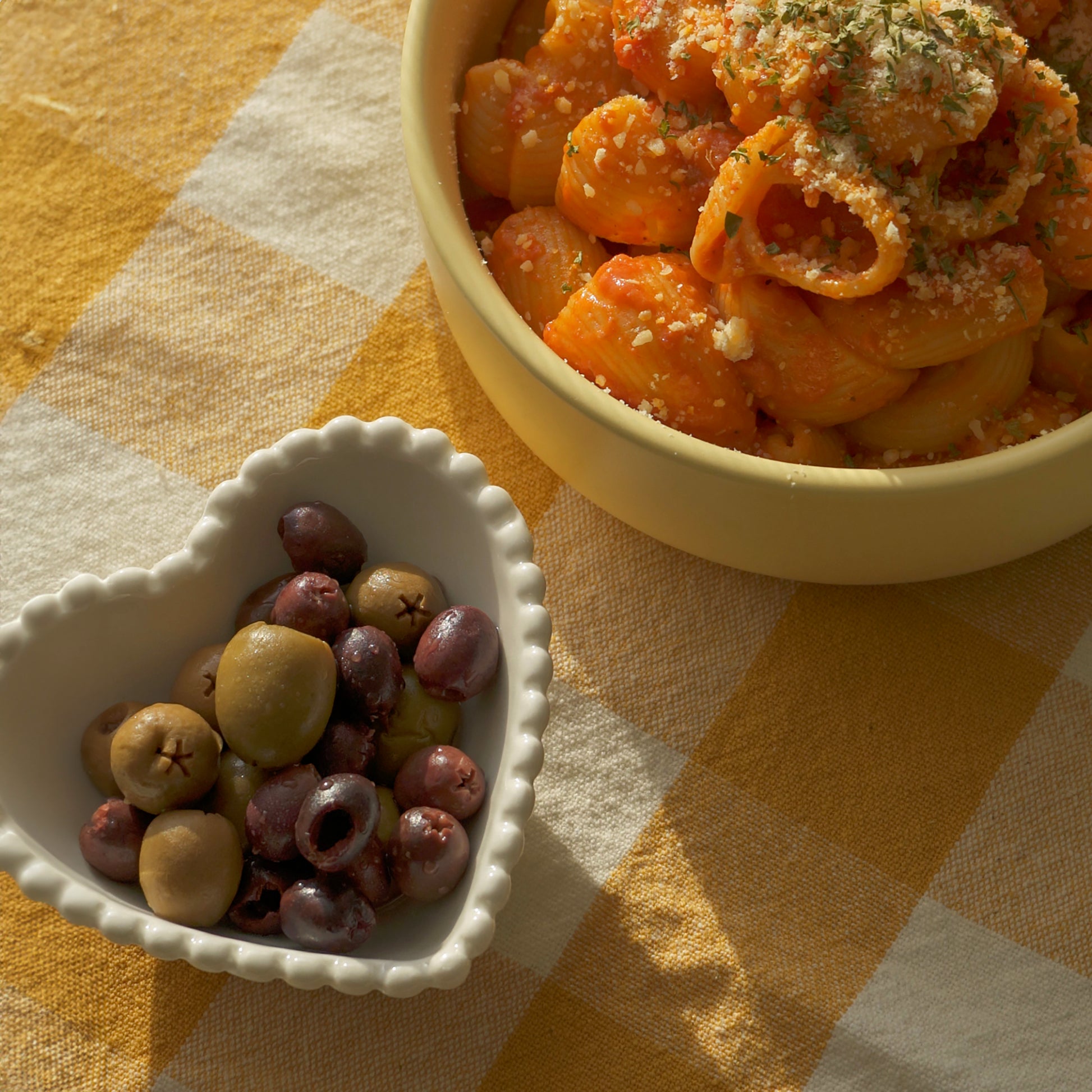 olives in a white ceramic beaded heart shaped bowl and lumache pasta in a yellow bowl placed on yellow checkerboard table cover