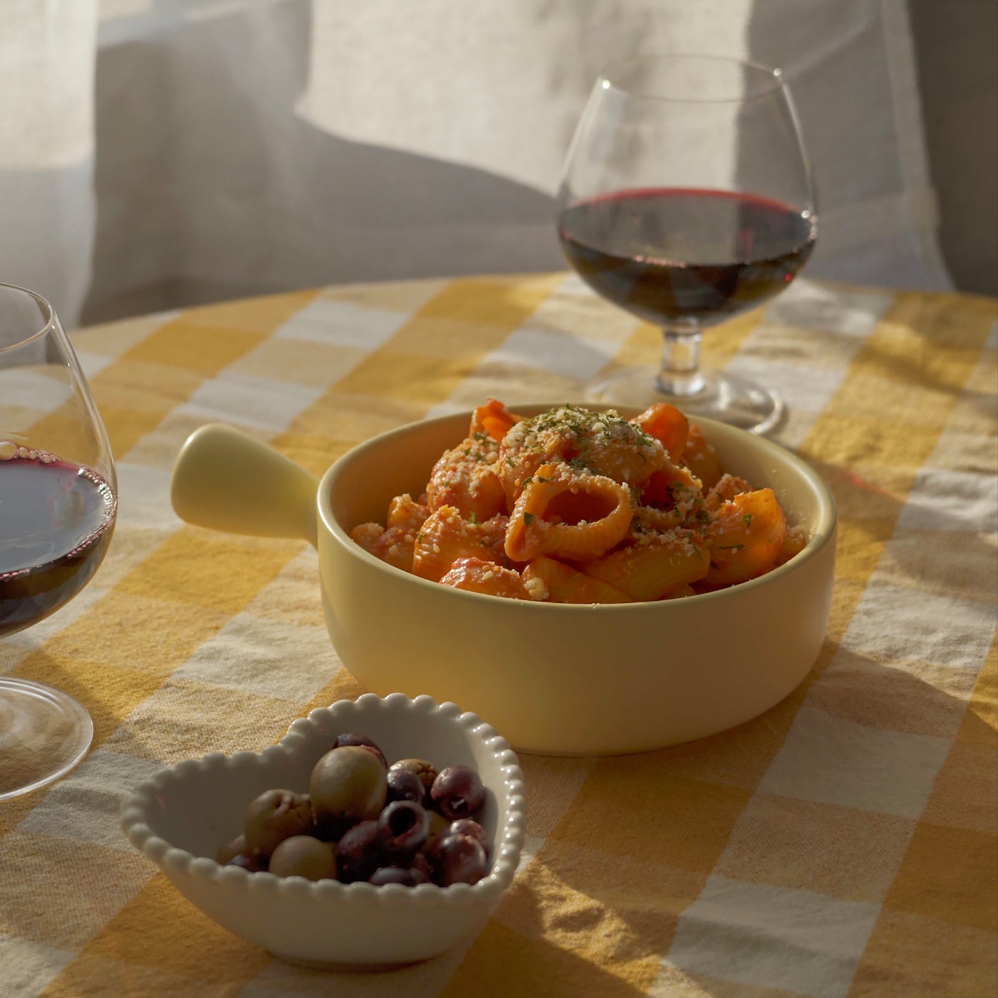 olives in a white ceramic beaded heart shaped bowl, lumache pasta in a yellow bowl, and two glassed of red wine placed on the table with yellow checkerboard table cover