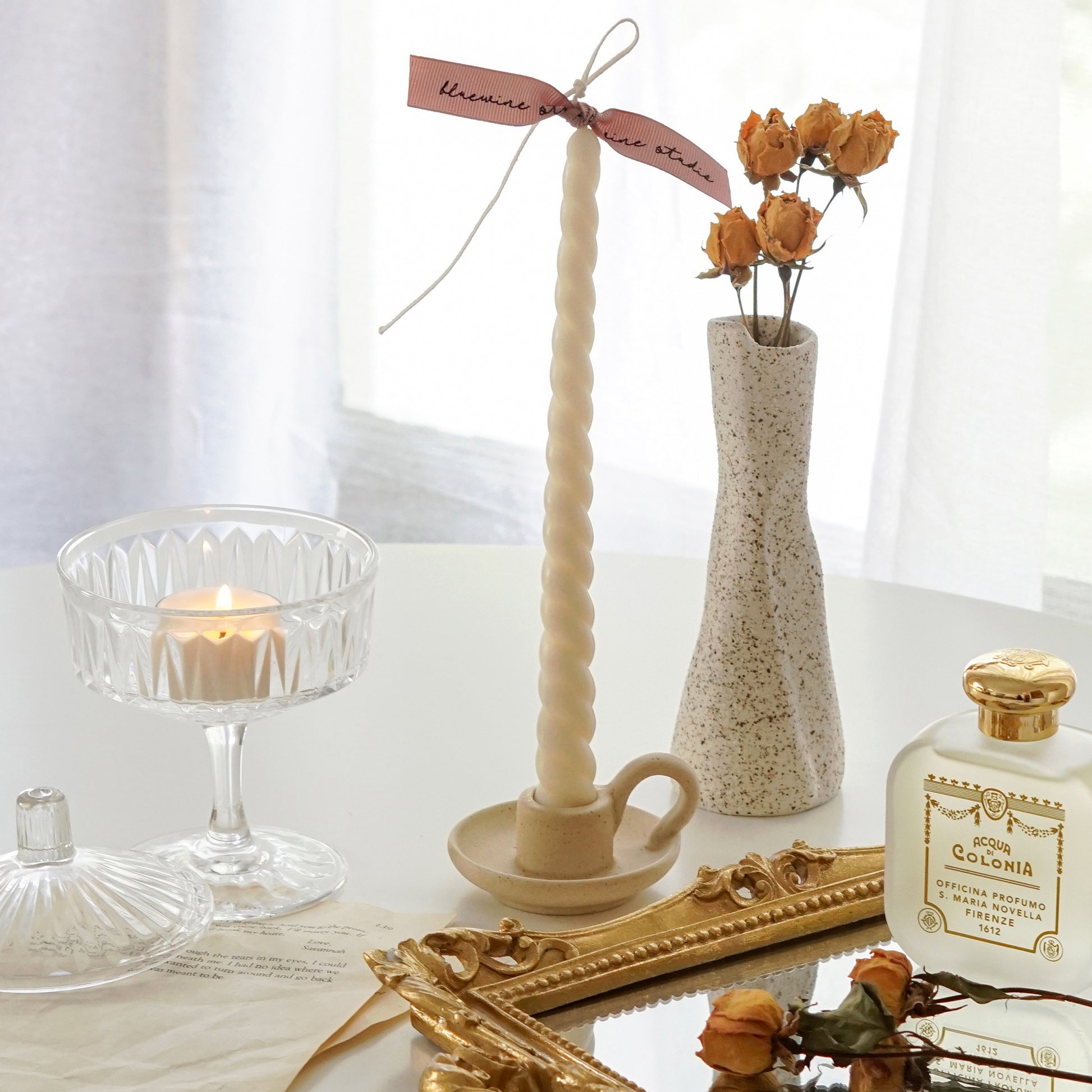 white twisted soy pillar candle with pink bluewine studio ribbon in a dotted beige ceramic taper candle holder, yellow dried roses in a ceramic dotted unique vase, a lit tealight candle in a coupe glass placed on book pages, and yellow rose dried flowers and santa maria novella perfume placed on rectangular gold french mirror tray on white table