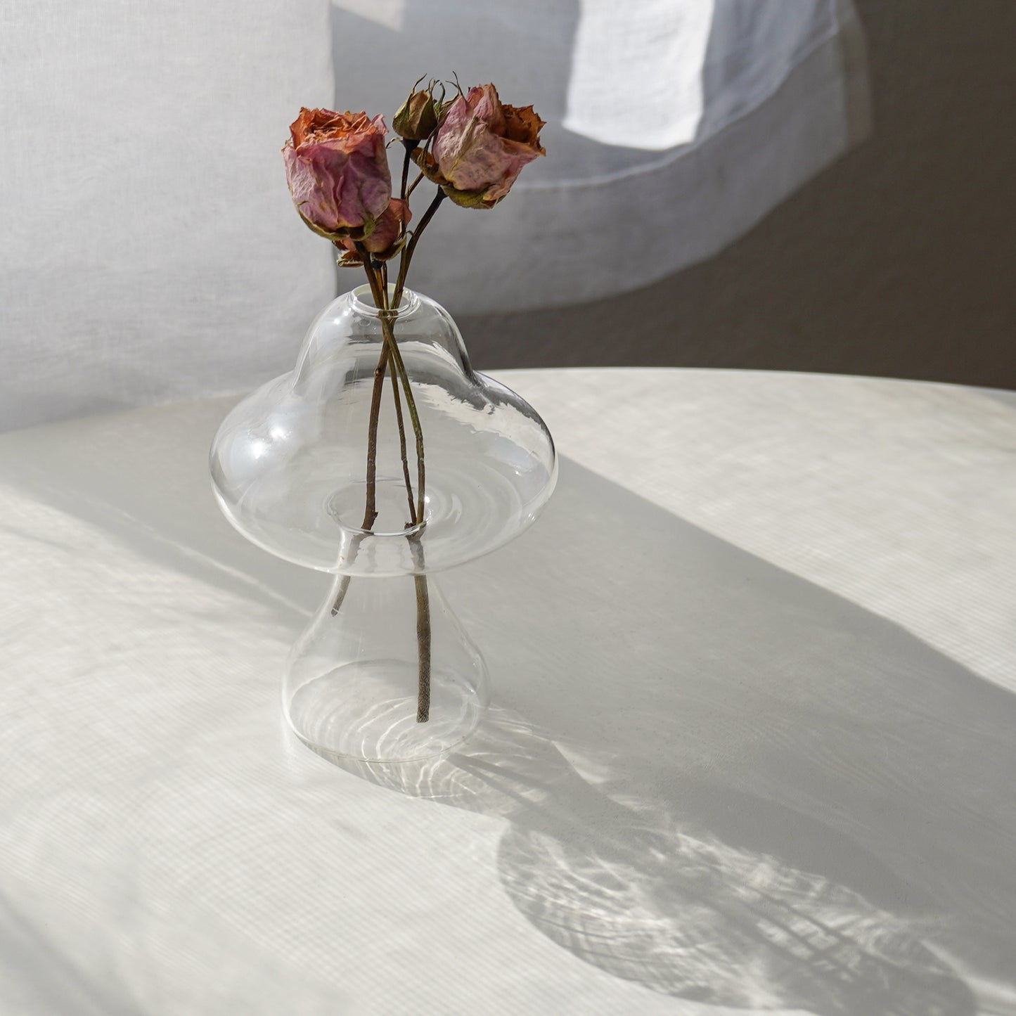 pink dried roses in a clear mushroom-shaped vase placed on a white round table by the window