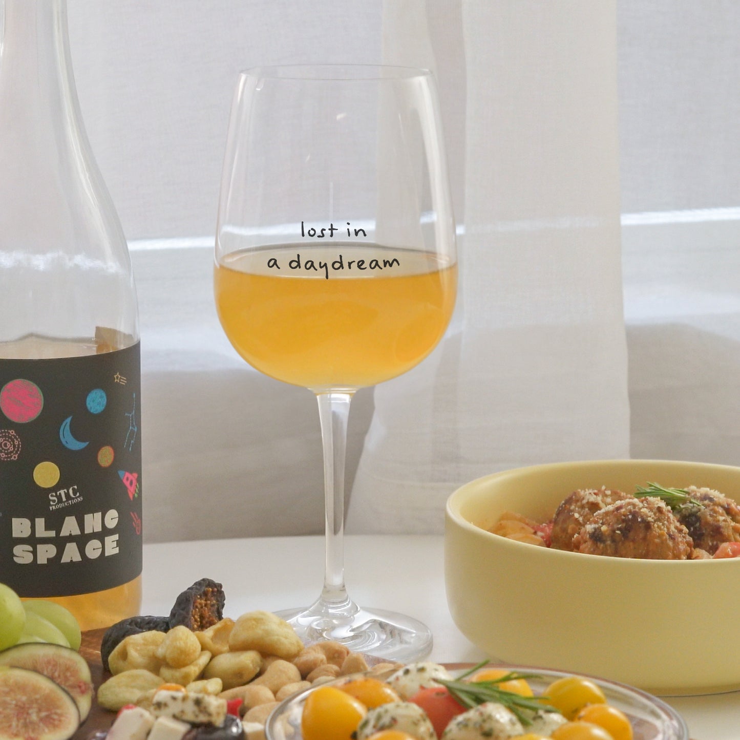 a glass of white wine in a glass printed with lost in a daydream, cheese board, meatballs, and a bottle of wine placed on a white round table