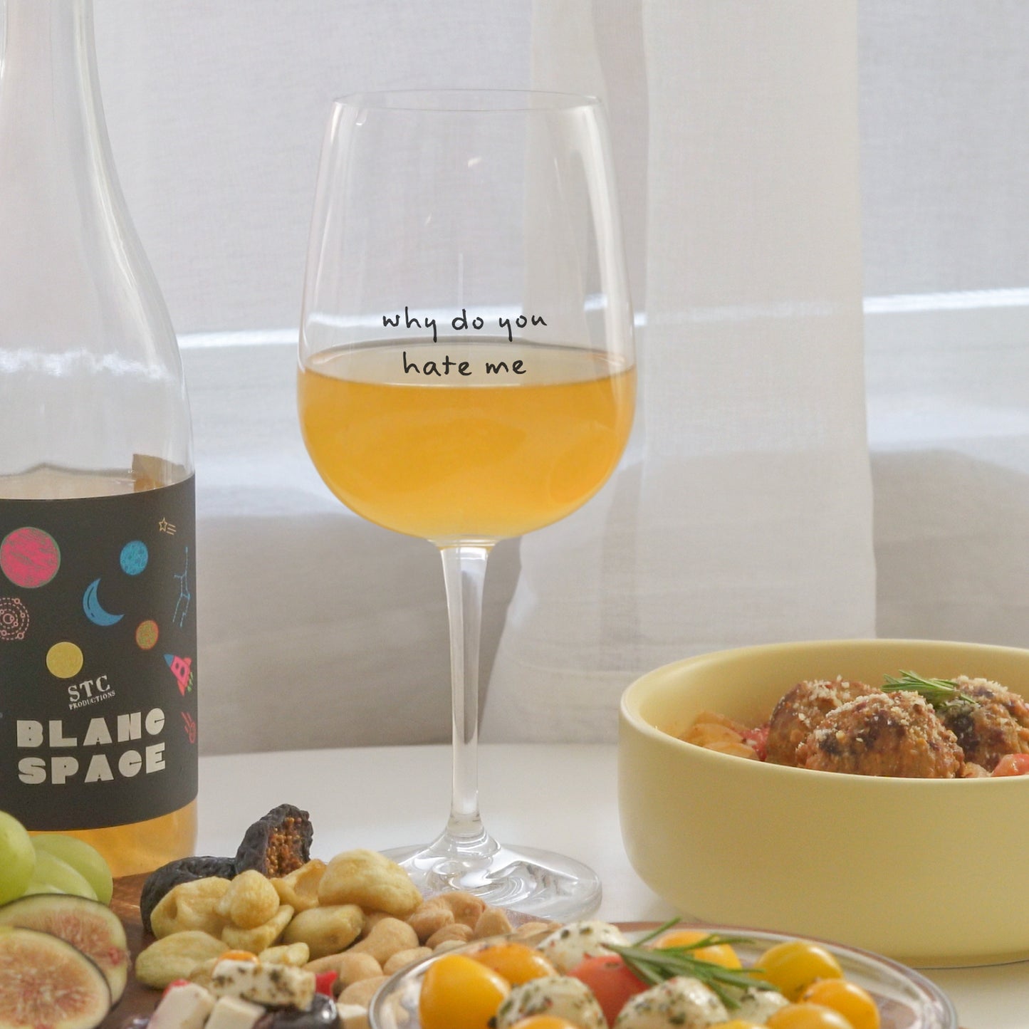 a glass of white wine in a glass printed with why do you hate me, cheese board, meatballs, and a bottle of wine placed on a white round table