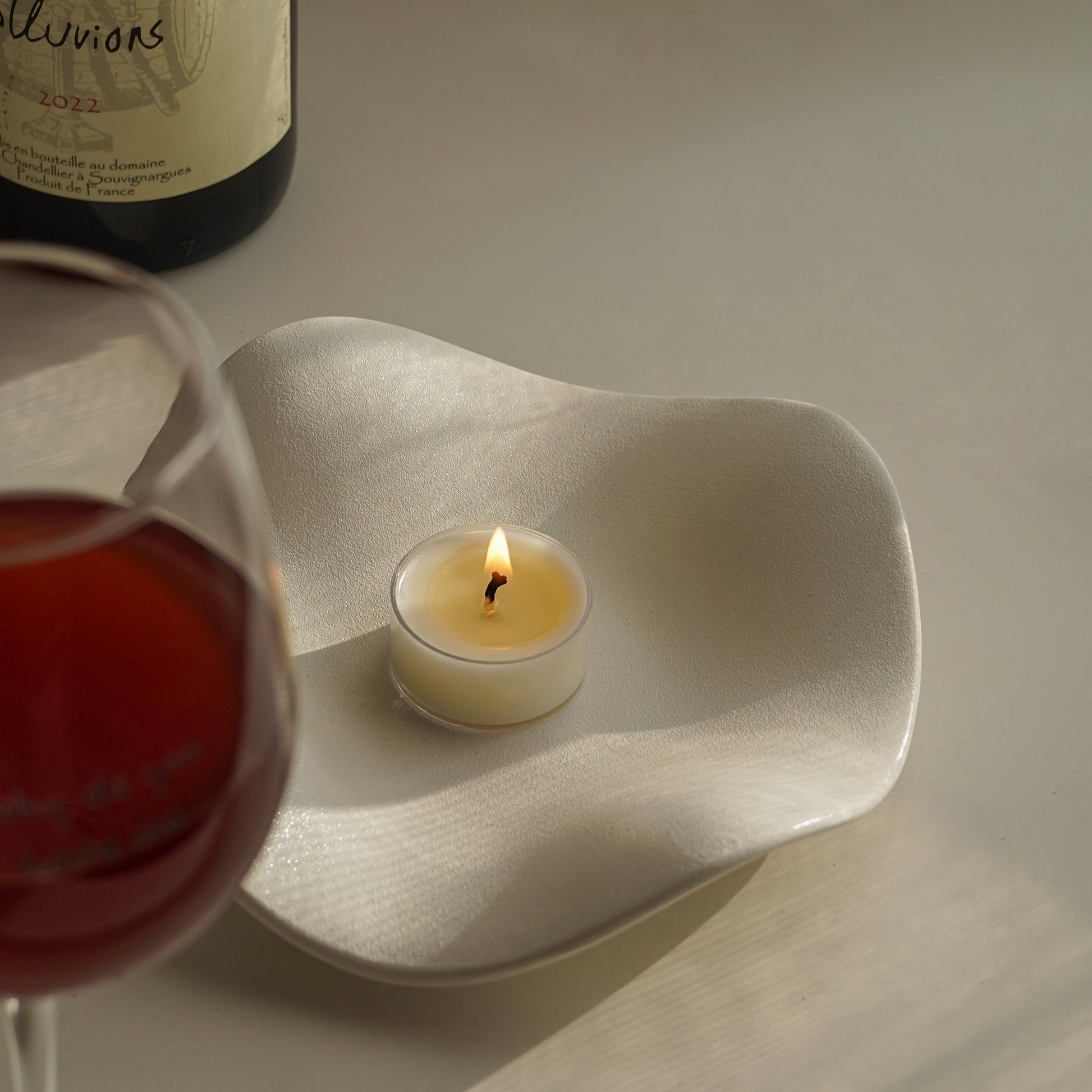 a glass of red wine, a bottle of red wine, and a lit tealight candle on a lotus flower shaped trinket dish placed on the table